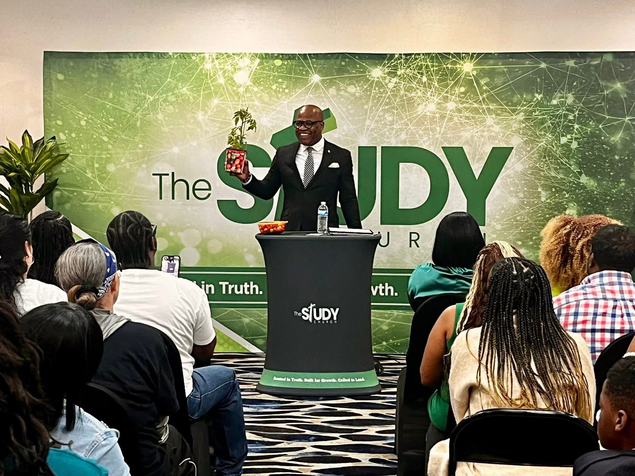 Man in suit speaking at a podium during a presentation, audience seated in front, green backdrop with text and graphics.