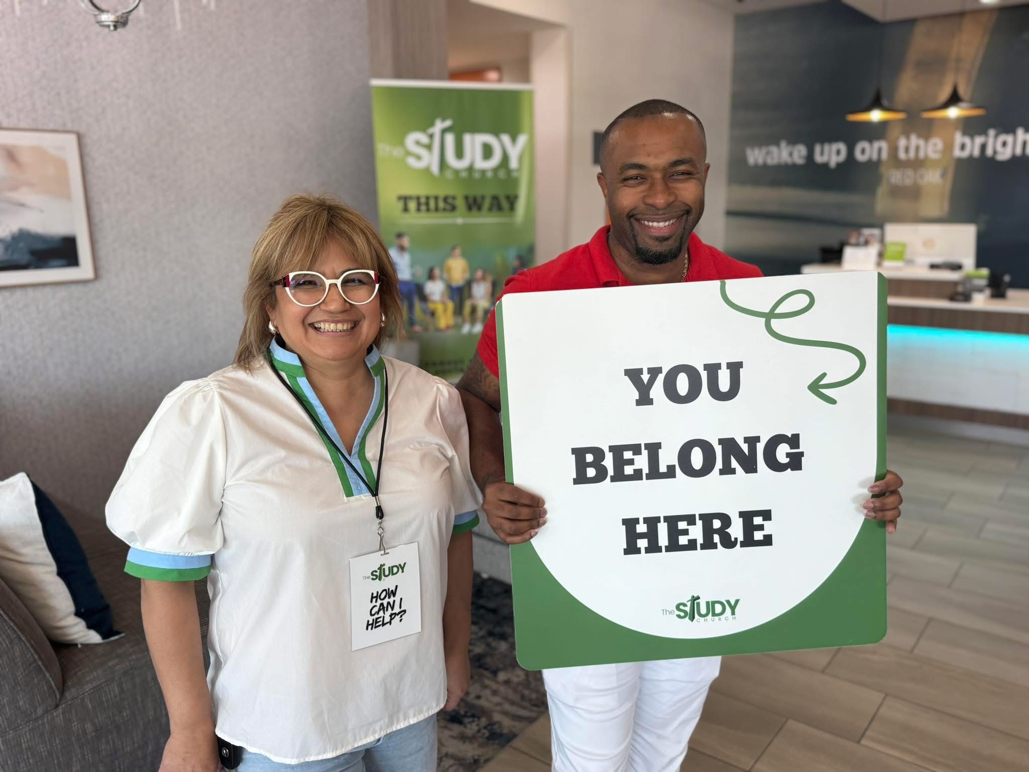 Two smiling people holding a sign that says 'You Belong Here' in a room with a study-related background.