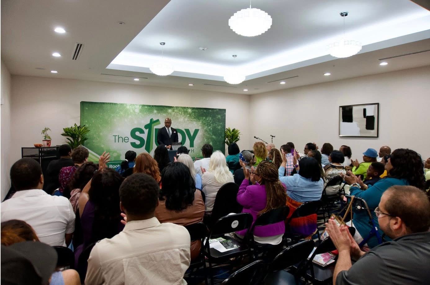 A speaker stands in front of a green banner reading 'The Study' in a conference room with an audience seated and listening.