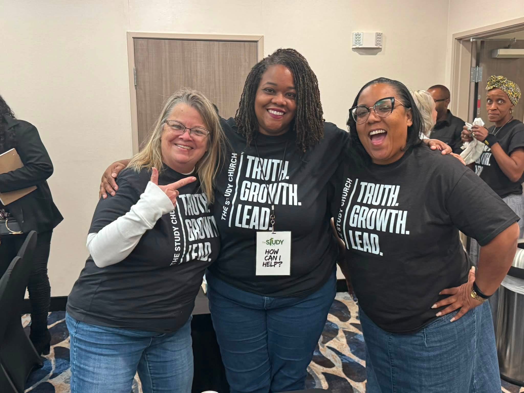 Three women smiling and posing together indoors, wearing matching black T-shirts with white text, with other people in the background.