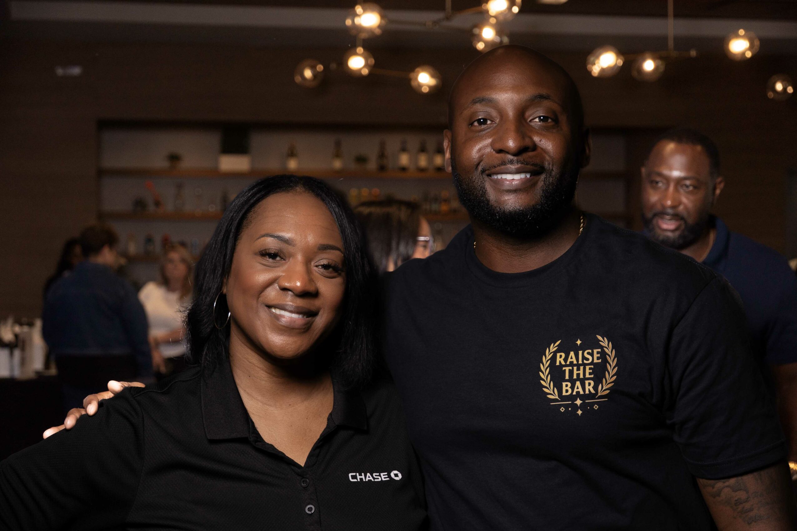 Two smiling people in a bar, one woman and one man, with warm lighting and other patrons in the background.