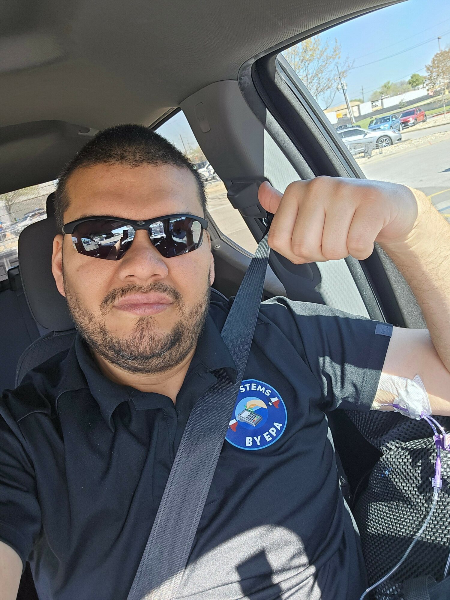 Man with sunglasses and a beard in a car, wearing a dark shirt with a patch, holding a strap, with a view outside the window.