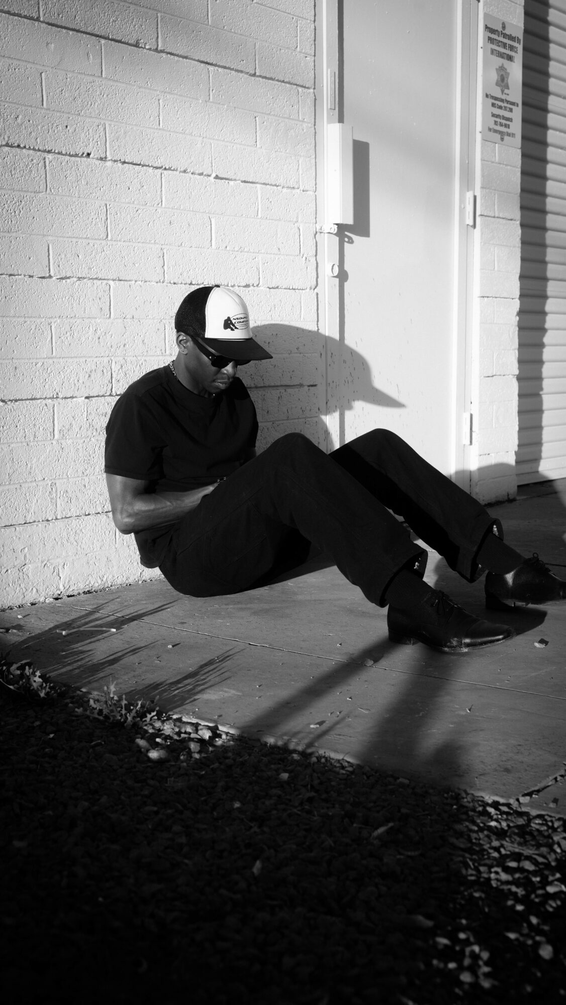 Person sitting on the ground against a brick wall, wearing a cap and dark clothing, with shadows cast on the sidewalk.