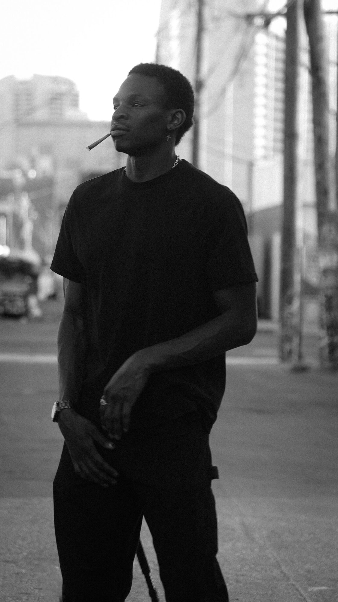 Young man standing outdoors in an urban setting, wearing a black t-shirt, with buildings and trees in the background.