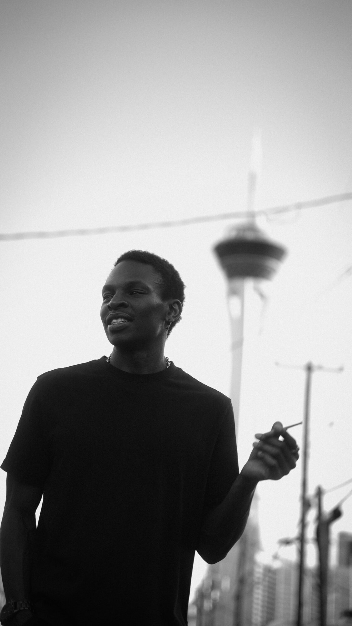 Young man holding a cigarette outdoors with a tower and power lines in the background.