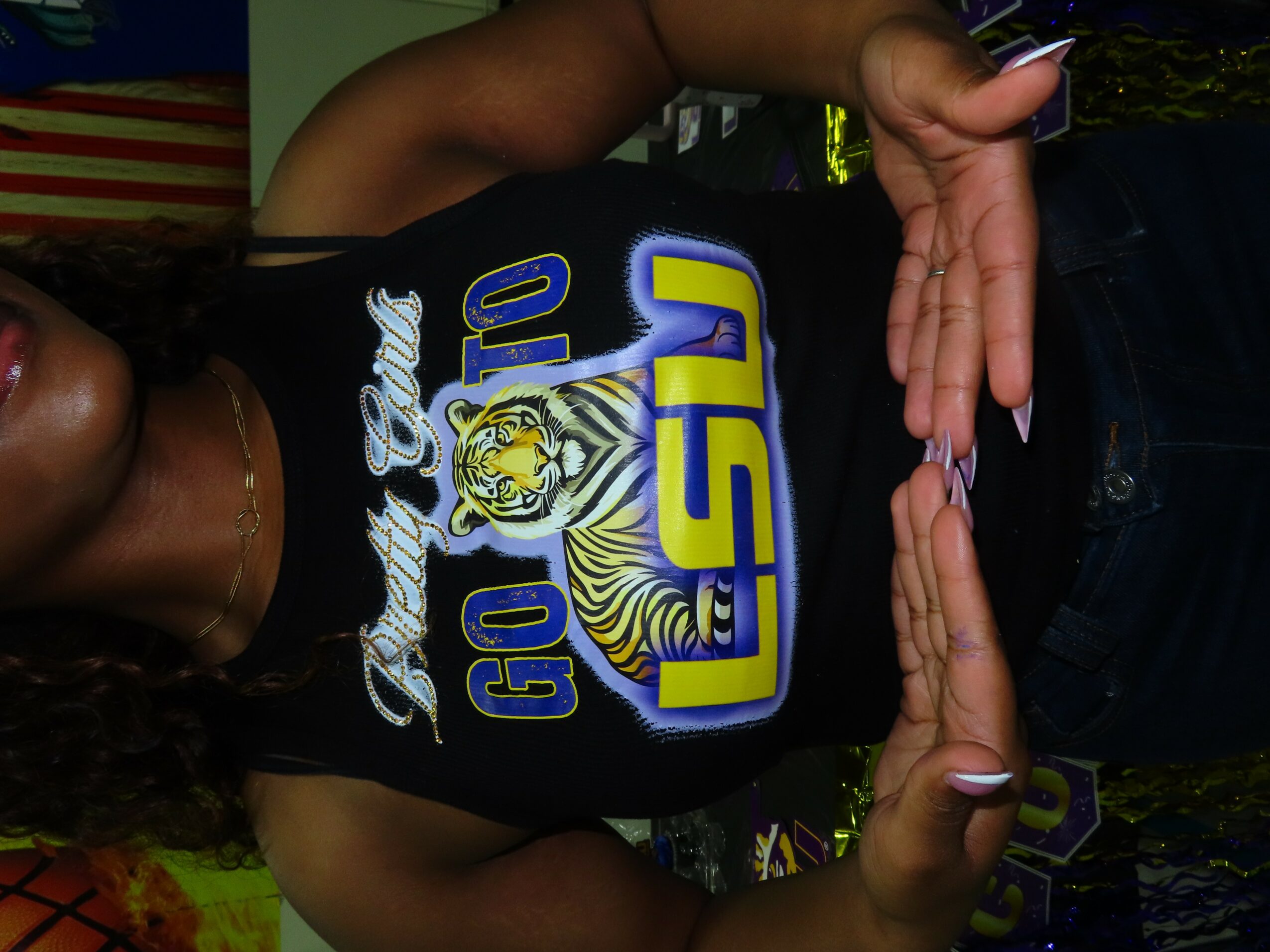 Person wearing a black LSU shirt with a tiger logo, making a heart shape with hands, indoors.
