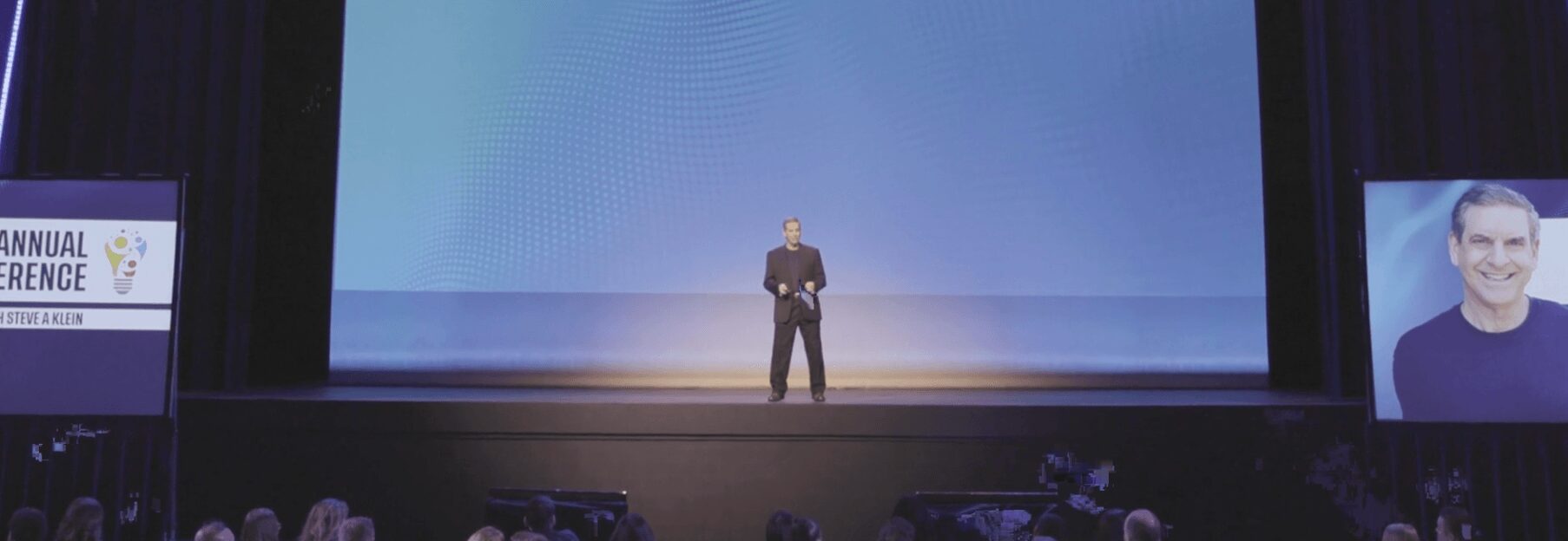 A person stands on a stage with a large screen behind, and a portrait of a smiling man on the right.