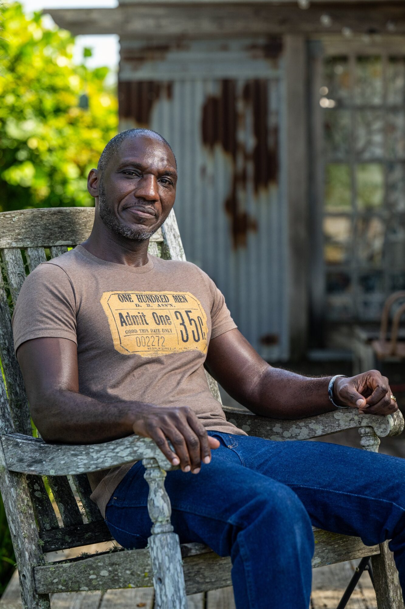 Man sitting outdoors on a wooden chair, wearing a brown T-shirt and blue jeans, with a rustic background.