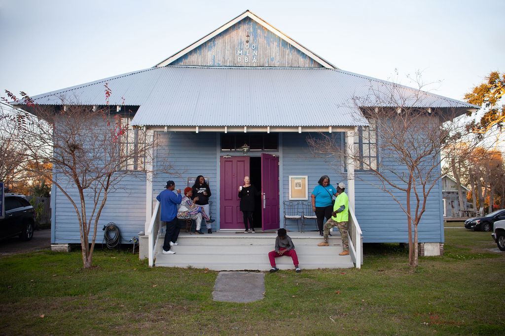 People gather on the front porch of a blue wooden building with a pink door, trees, and a grassy lawn.