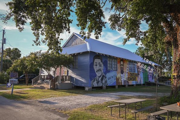 Building with murals of people and colorful artwork, surrounded by trees and outdoor seating, under a partly cloudy sky.