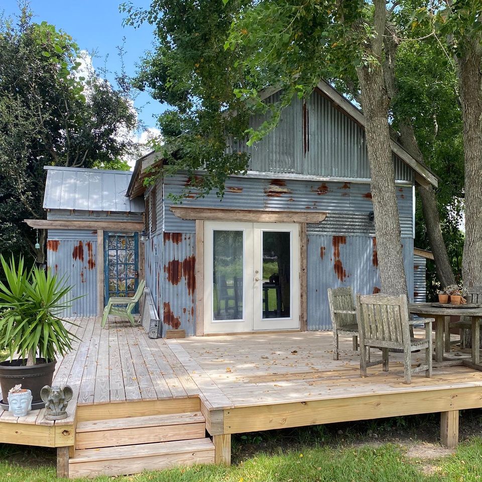 Rusty metal house with a wooden deck, surrounded by trees and outdoor furniture, under a partly cloudy sky.