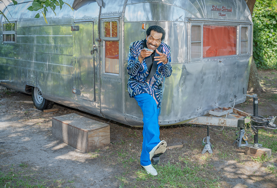 Person standing in front of a shiny metal trailer, smiling, wearing a patterned shirt and blue pants, outdoors.