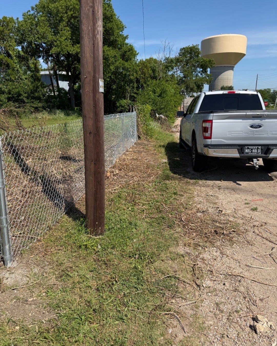 A white pickup truck parked on a dirt area next to a chain-link fence and a utility pole, with a water tower in the background.