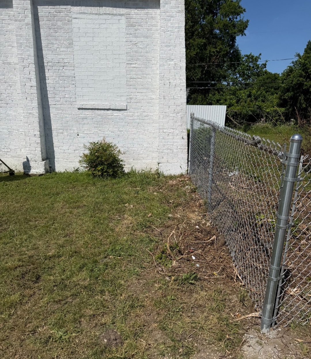 White brick building with a small bush, chain-link fence, and grassy yard under a clear blue sky.