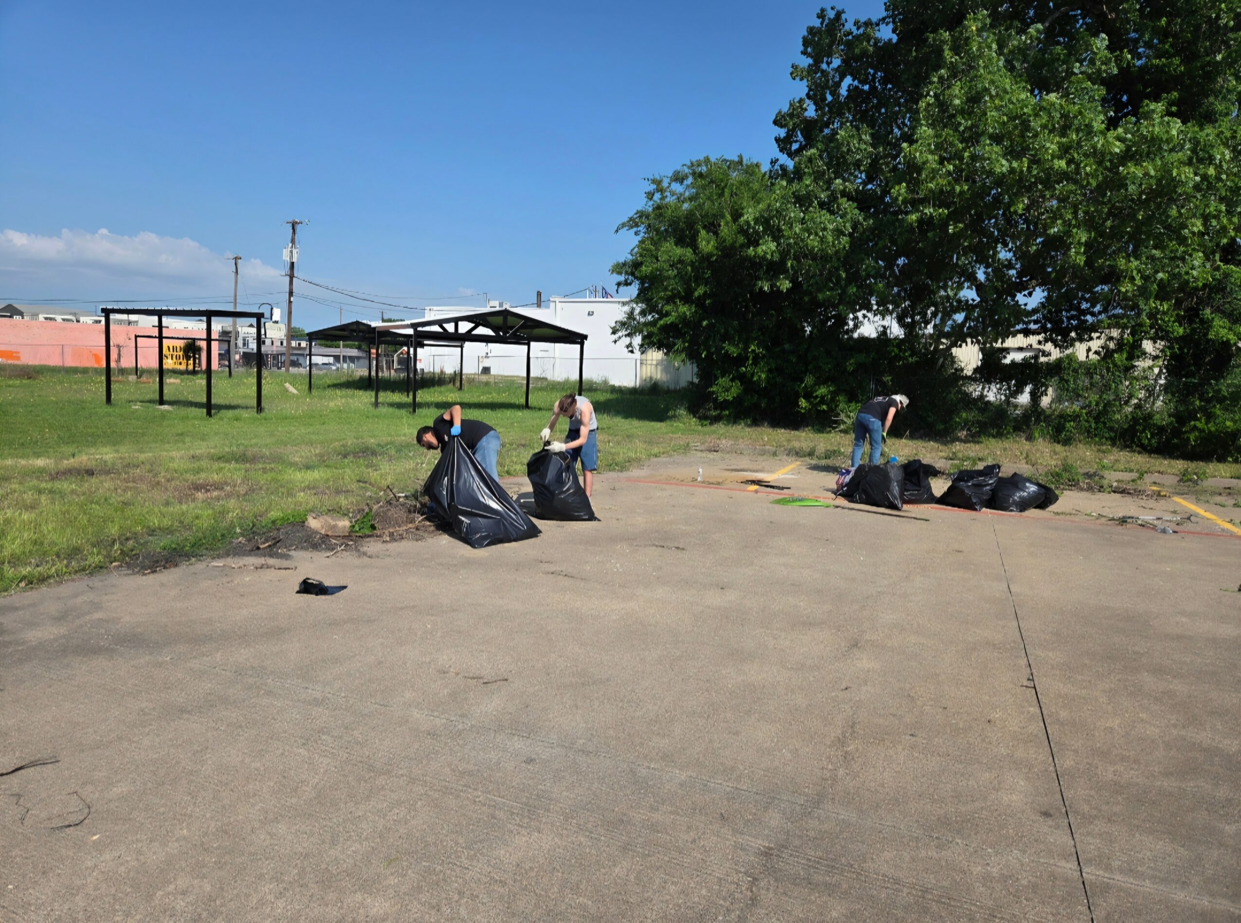Three people are cleaning up trash in an open outdoor area with grass, trees, and a building in the background.