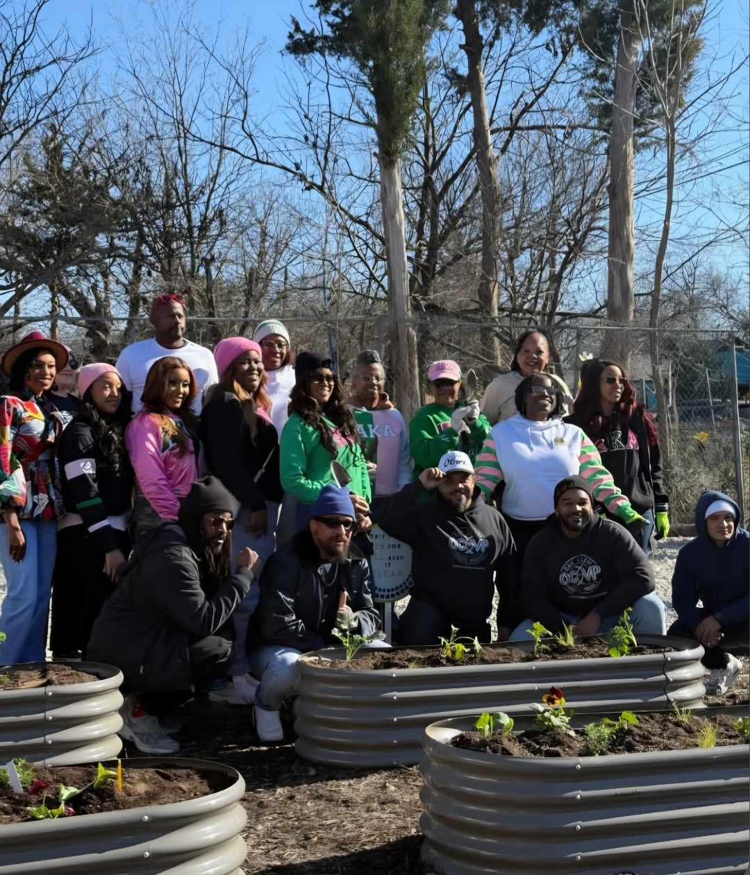 Group of people outdoors in a garden, some standing and some kneeling, with trees and a fence in the background.