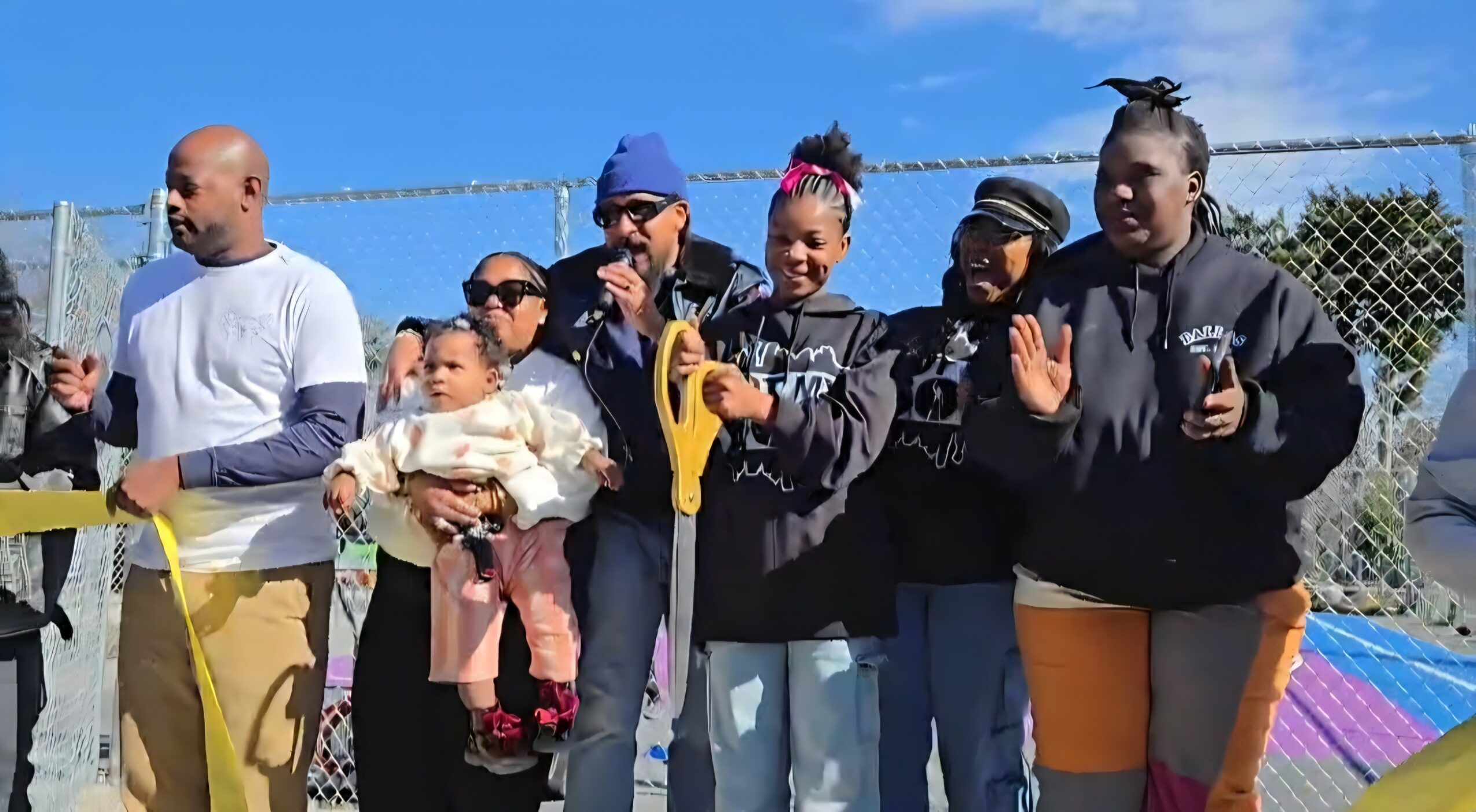 Group of people standing outdoors near a chain-link fence, some clapping and holding objects, with a clear sky background.