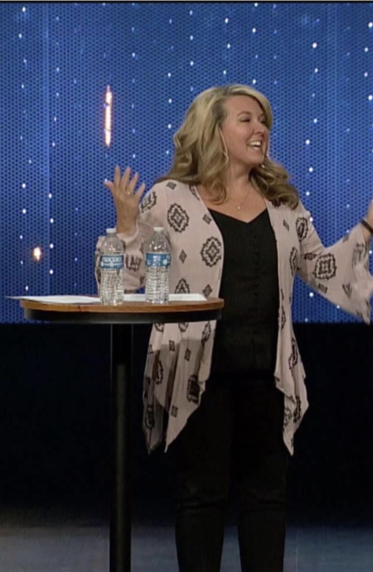 Woman smiling and gesturing with hands on stage with a starry background, standing next to a table with water bottles.