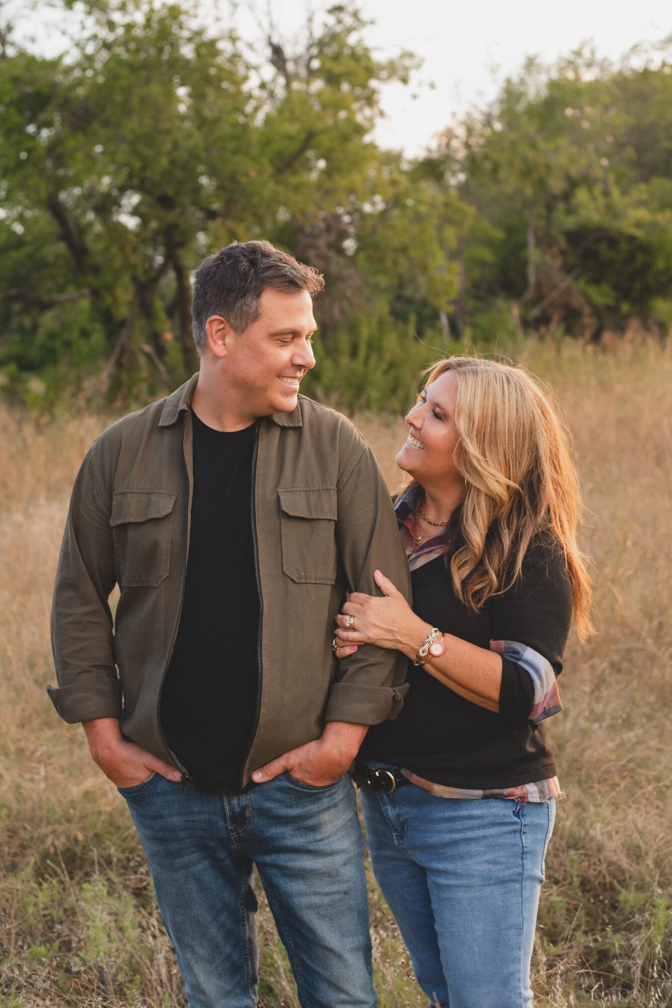 A man and woman standing outdoors in a grassy area with trees, smiling at each other.