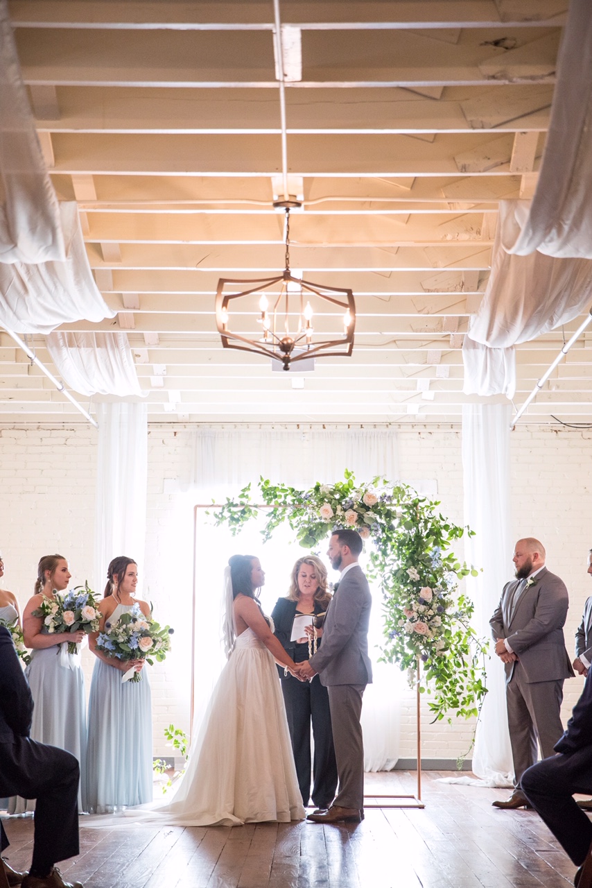 Wedding ceremony with couple holding hands, officiant, bridesmaids, groomsman, and guests in a bright room with large window and greenery.