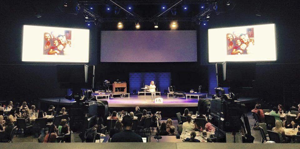 Large stage with two screens displaying images, audience seated in front, stage lighting, and equipment visible.