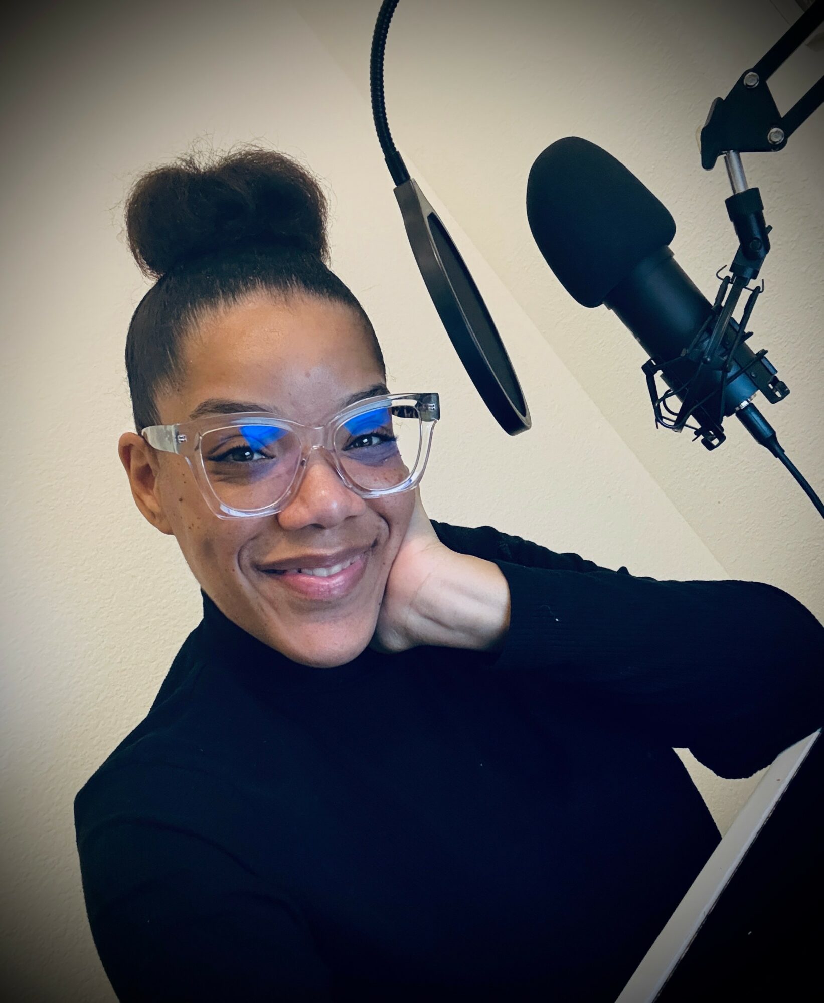 Woman with glasses smiling, sitting in front of a microphone, with a pop filter and headphones, in a recording studio.