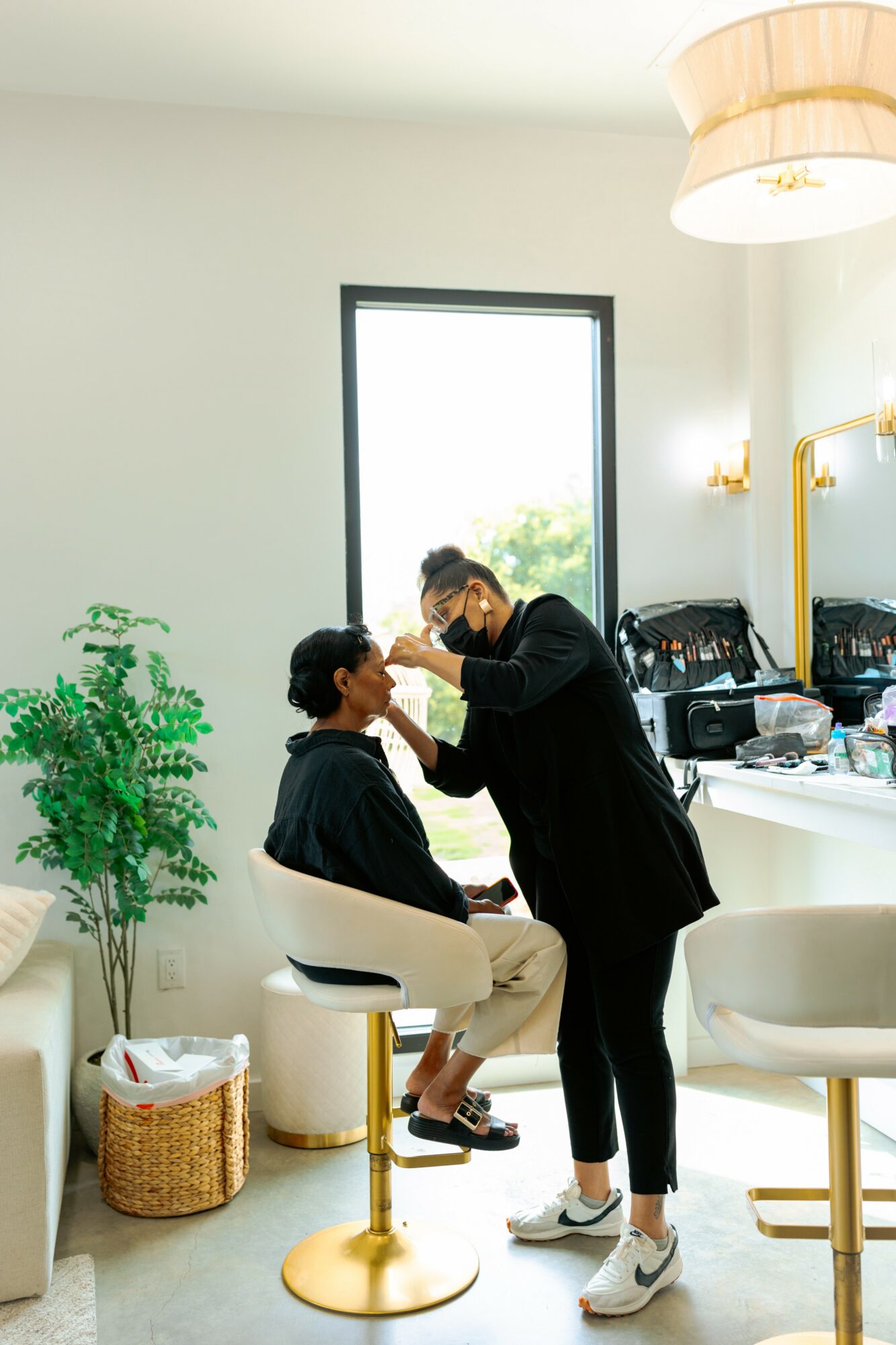 Person applying makeup to another person seated in a bright room with a large window, plants, and modern decor.