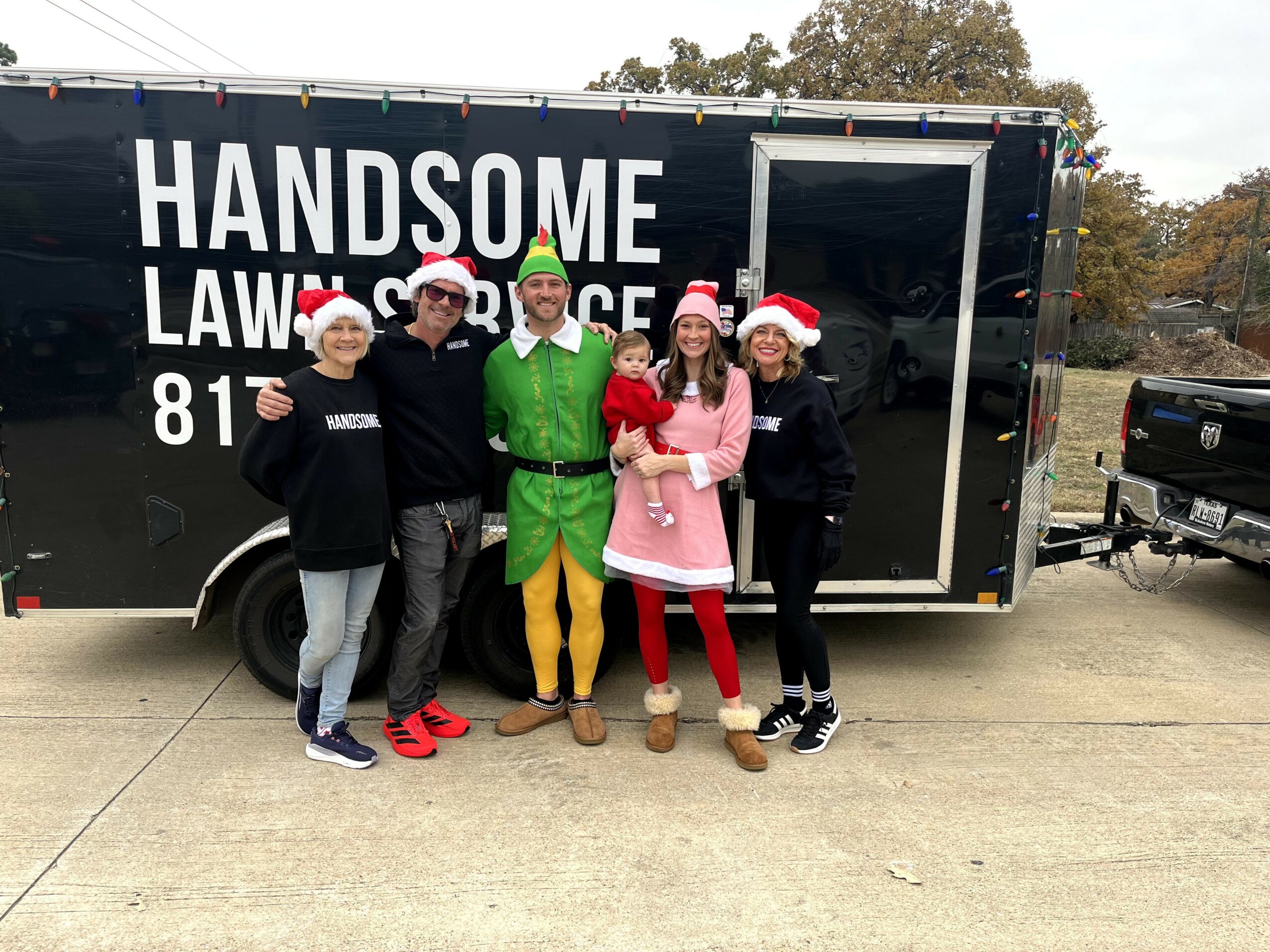 Group of five people wearing Santa hats standing in front of a black trailer with white text, outdoors on a concrete surface.