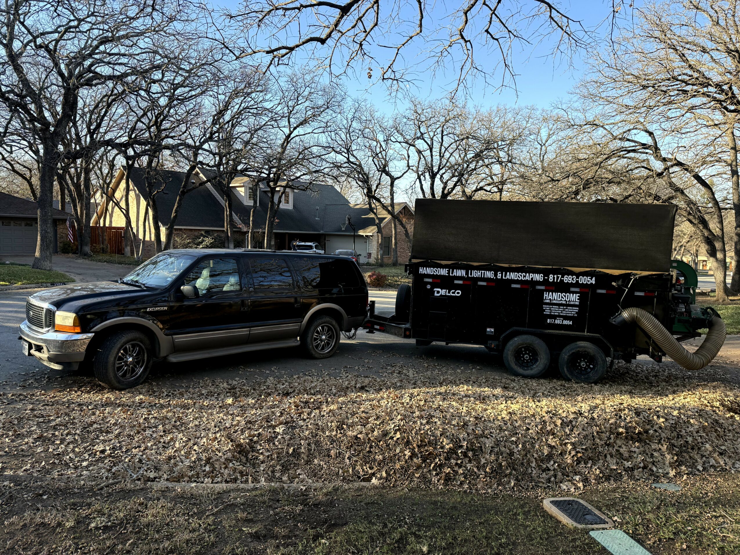 Black pickup truck connected to a black trailer with a cover, parked on a gravel surface with trees and houses in the background.