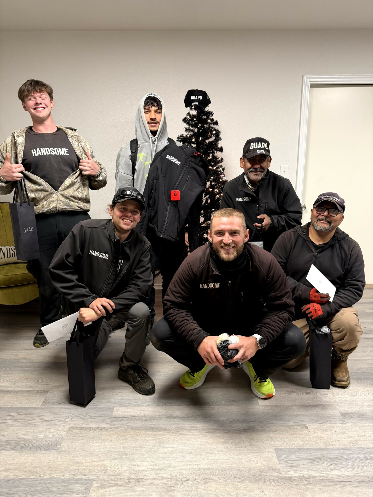Group of seven people posing indoors with a small Christmas tree, some holding papers, smiling, in casual clothing, in front of a white wall.