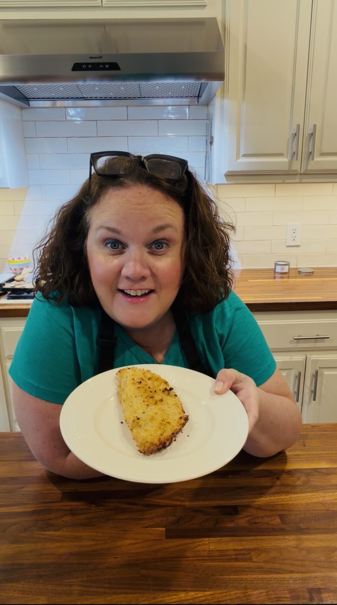 Woman with curly hair holding a plate with a piece of food, smiling in a kitchen, wearing glasses on her head, green shirt.
