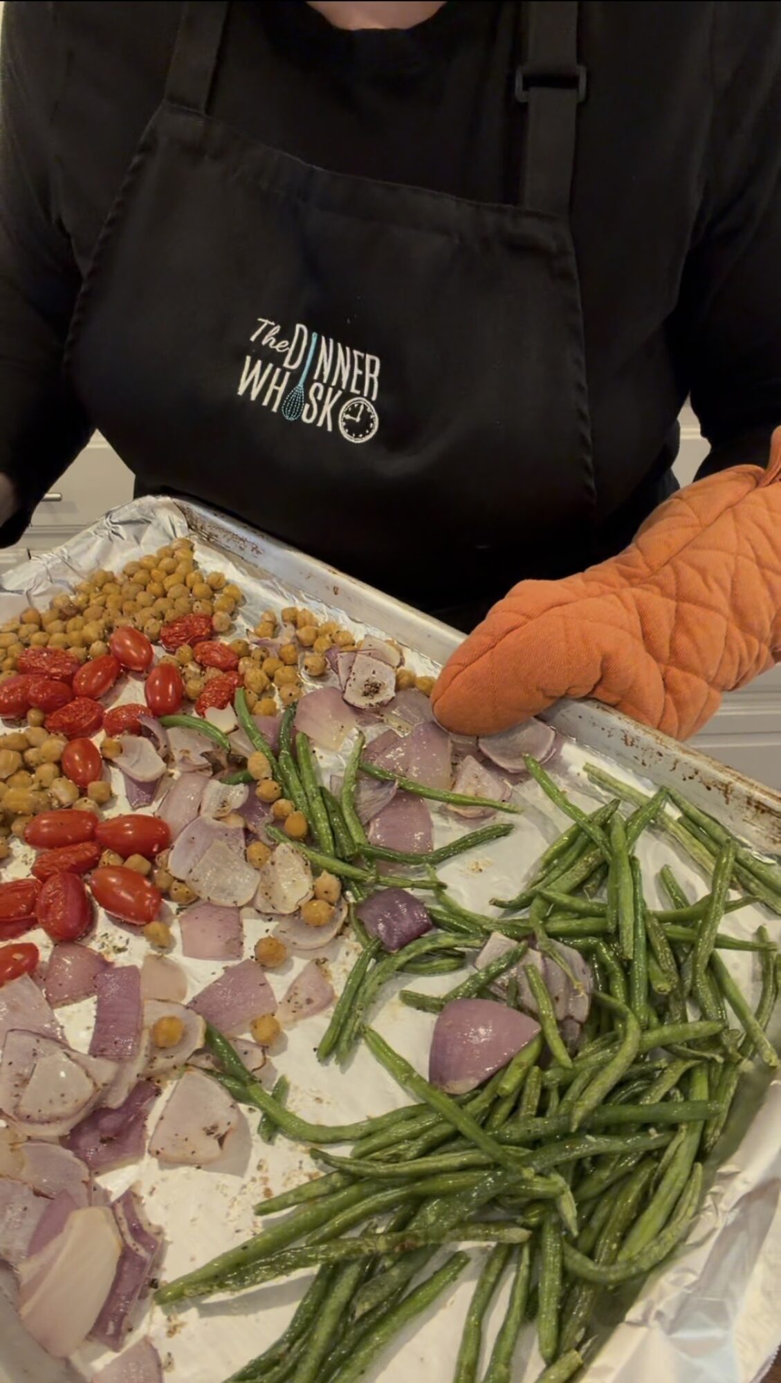 Person wearing a black apron with white text, preparing a tray of chopped vegetables including green beans, onions, and cherry tomatoes.