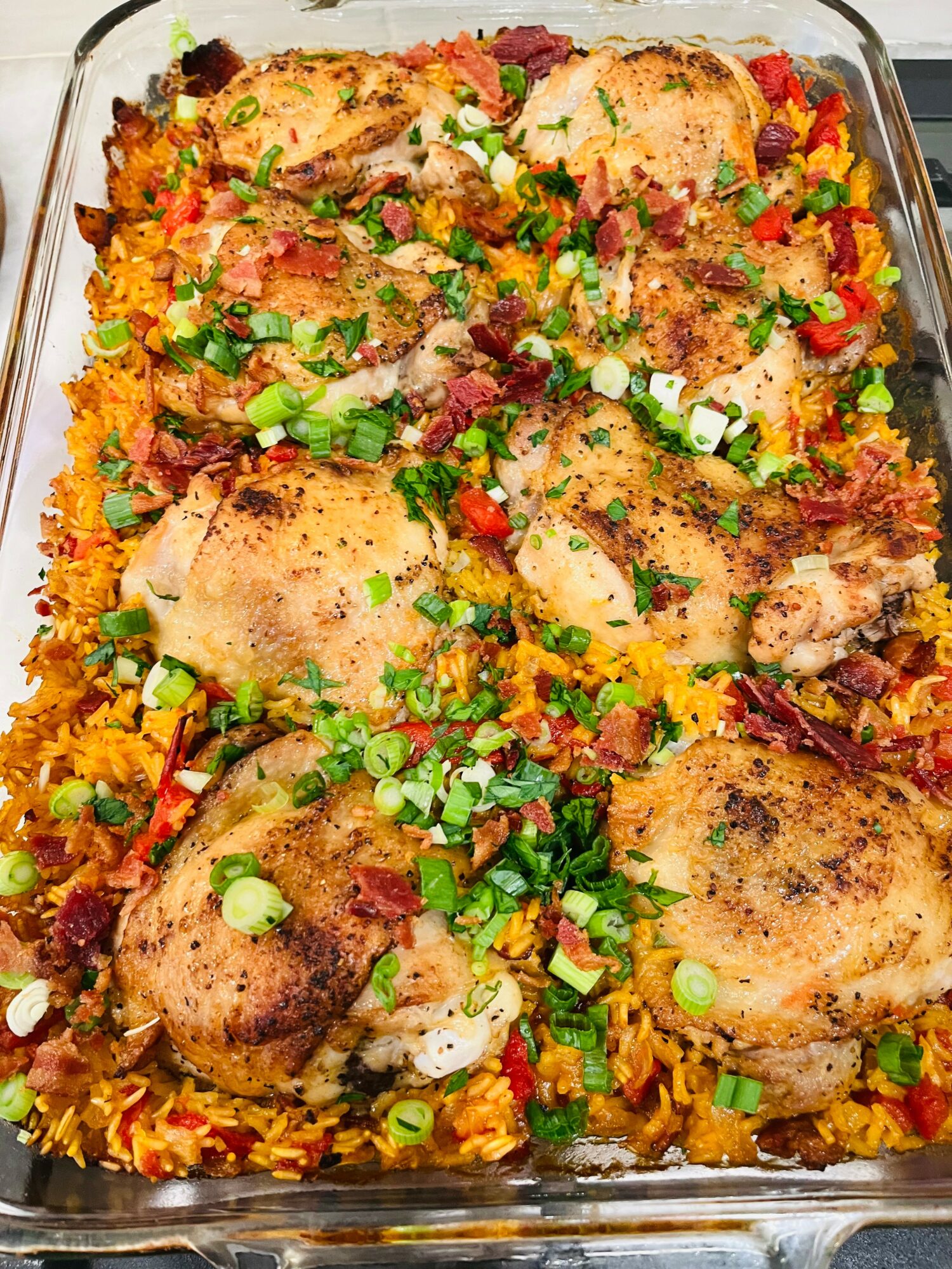 Baked chicken thighs on seasoned rice with chopped green onions and red peppers in a glass dish.