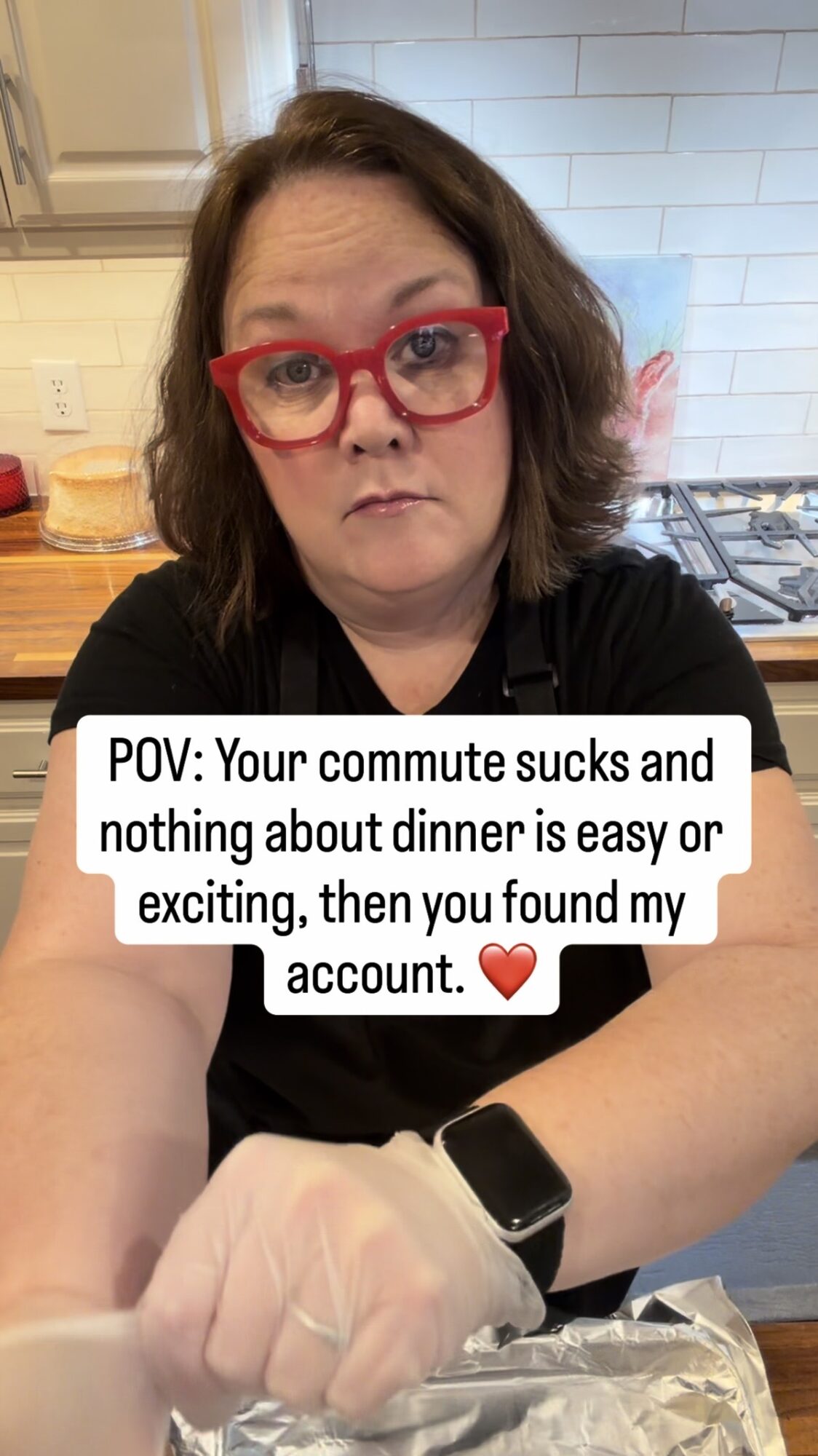 Woman with red glasses in a kitchen, wearing a black shirt and a smartwatch, looking at the camera with a serious expression.