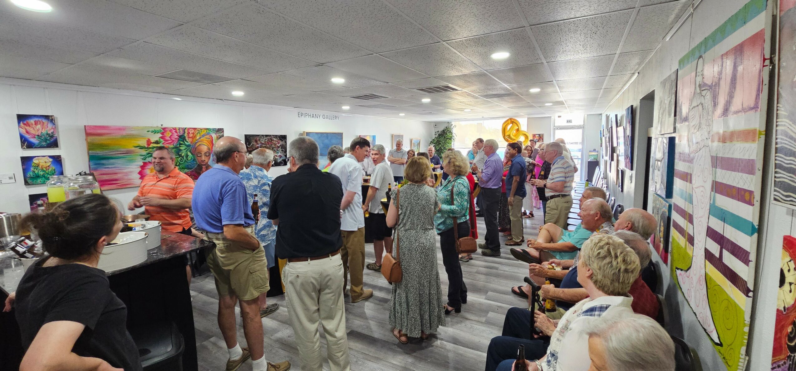 People gather in an art gallery with colorful paintings on the walls, some seated and others standing, engaging with artwork.