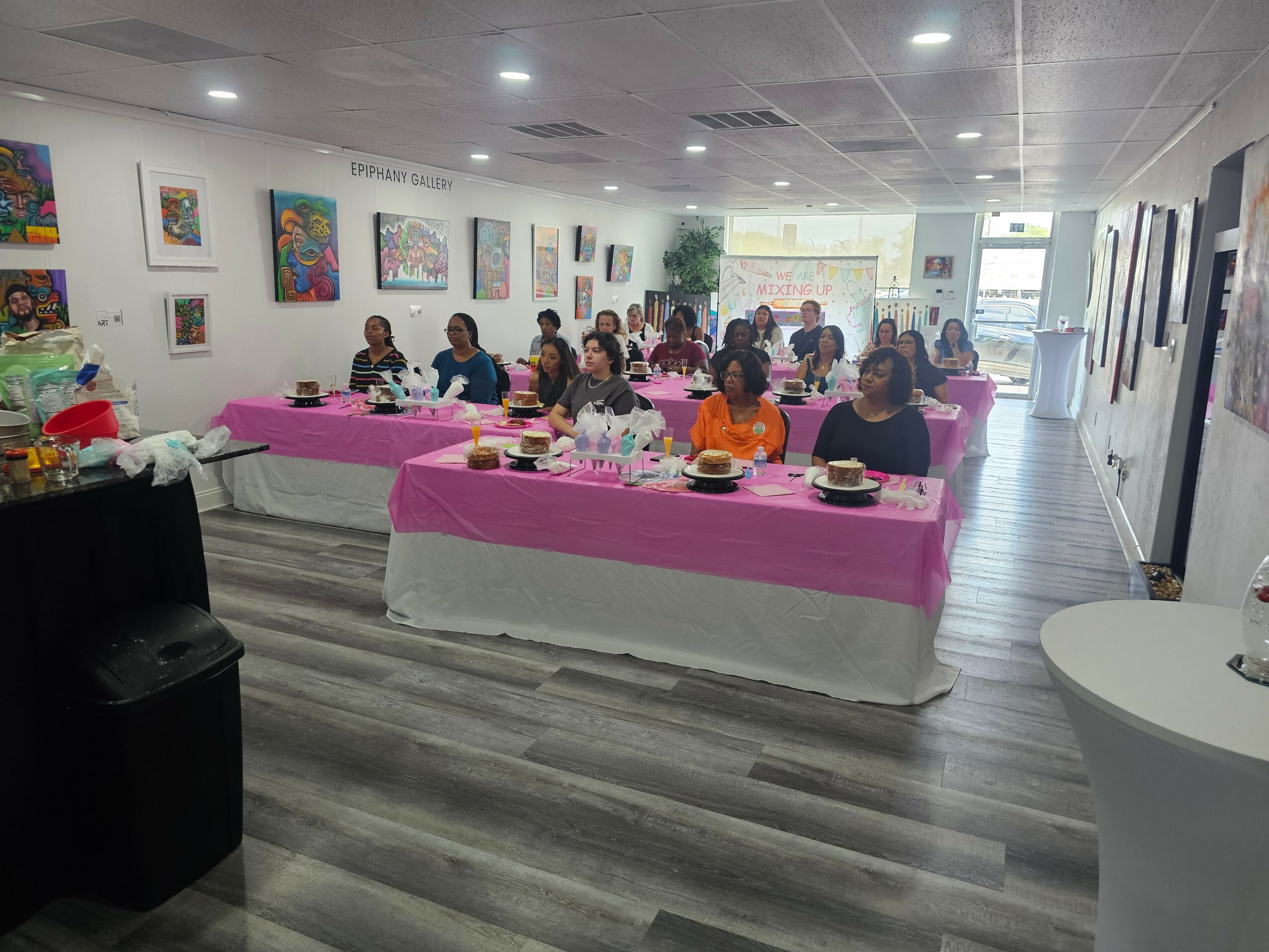 People gathered around tables with pink tablecloths in a brightly lit art gallery, artwork displayed on walls, and a window at the back.