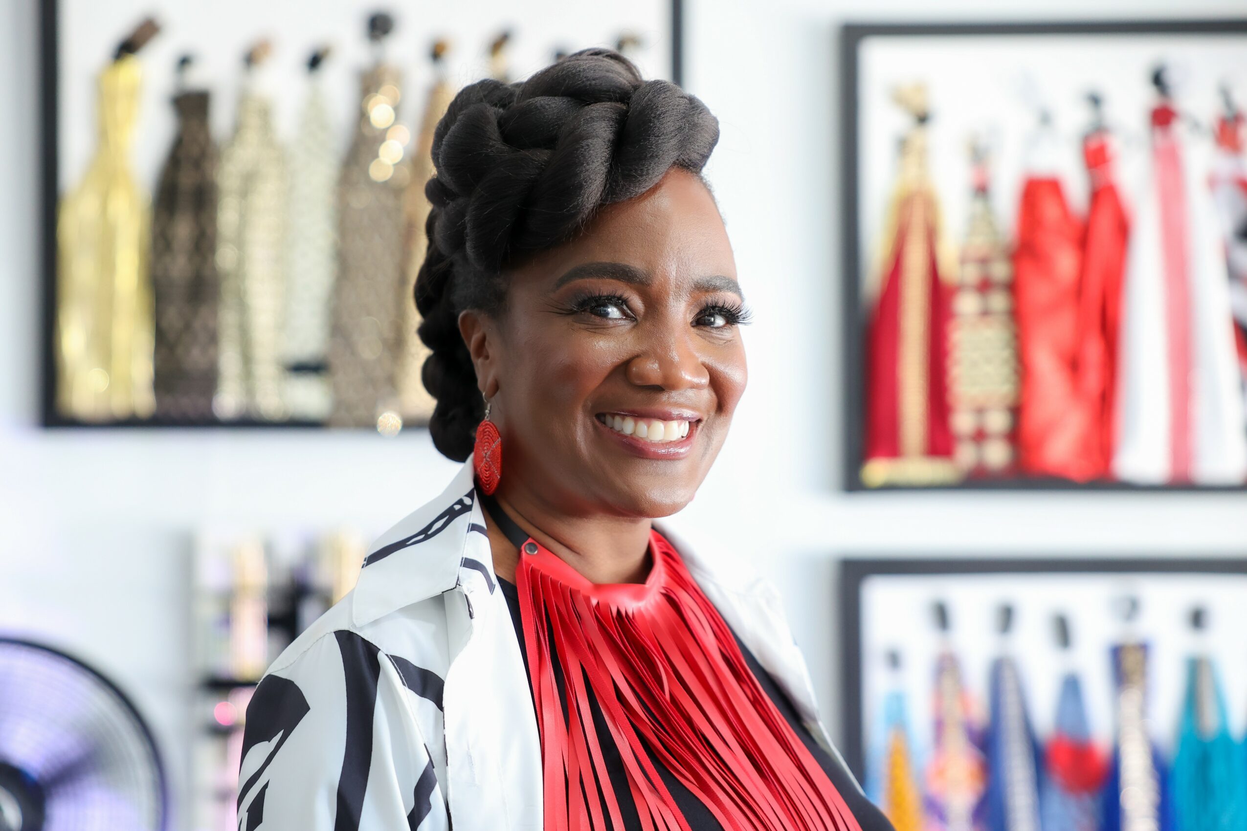 Smiling woman with styled hair and red earrings in front of clothing displays, wearing a patterned jacket and red top.