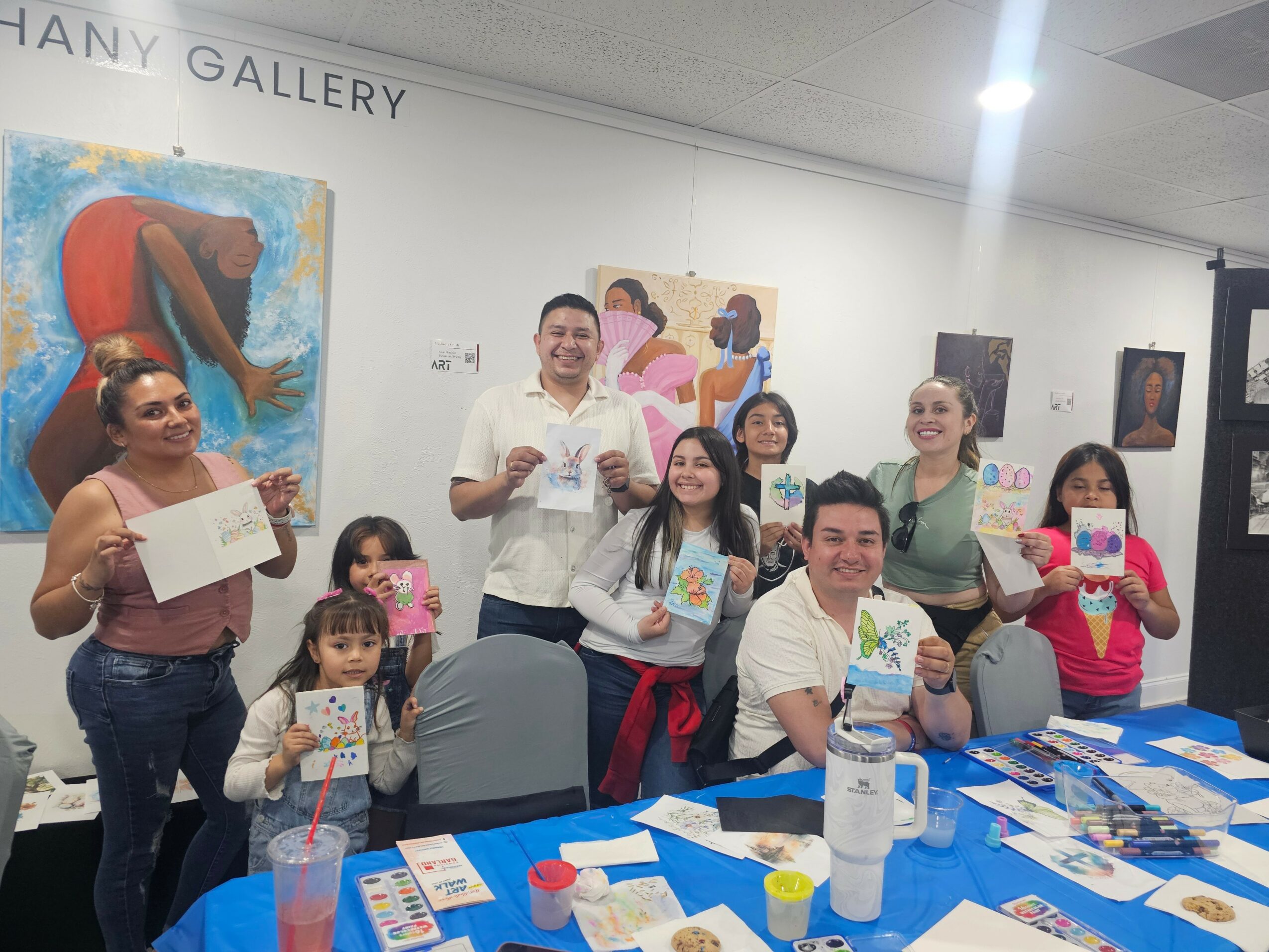 Group of people at an art gallery holding artwork, smiling, with paintings on the wall behind them.