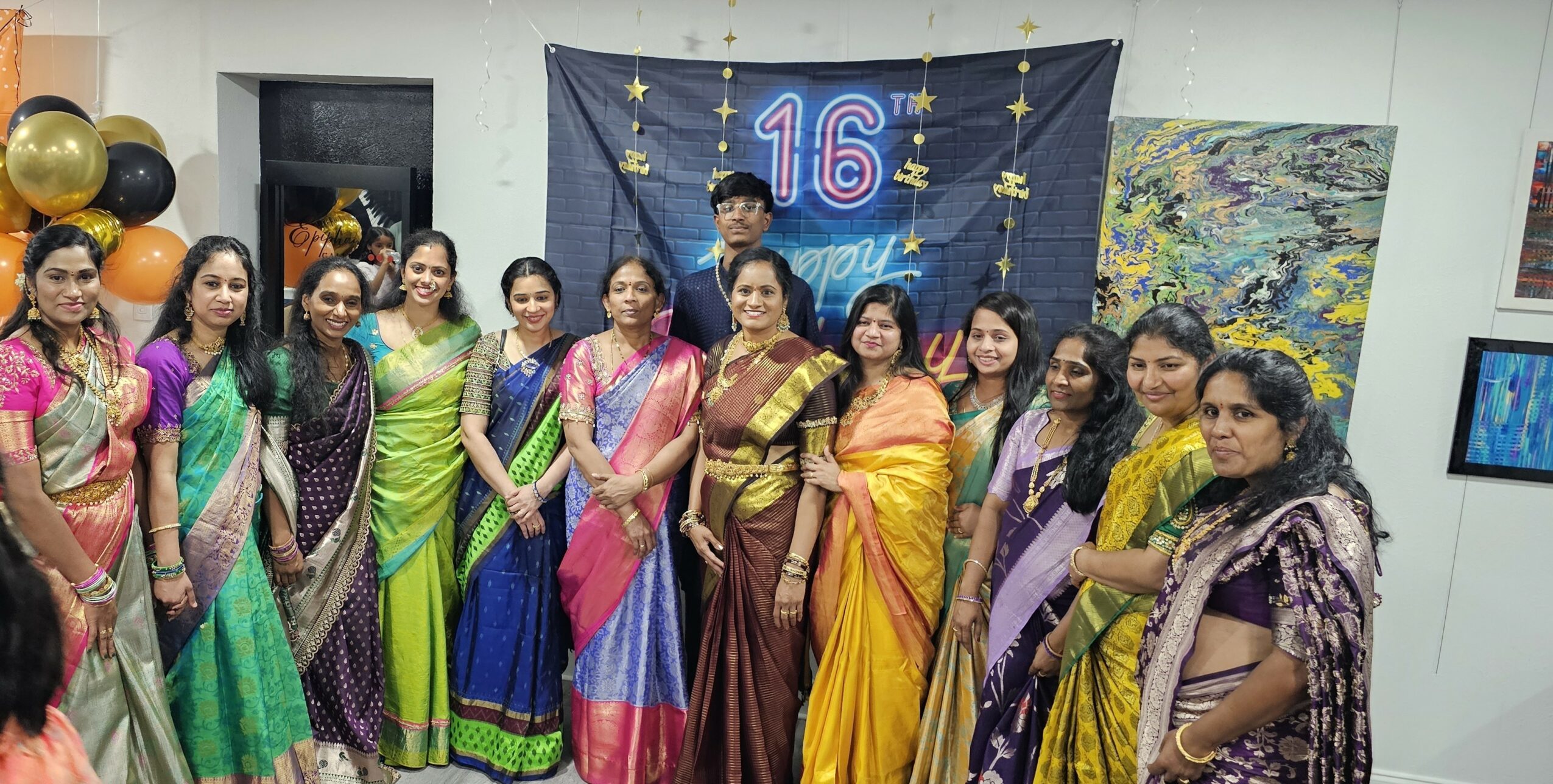 Group of women in colorful traditional dresses standing in front of a decorated backdrop with the number 16 and stars.