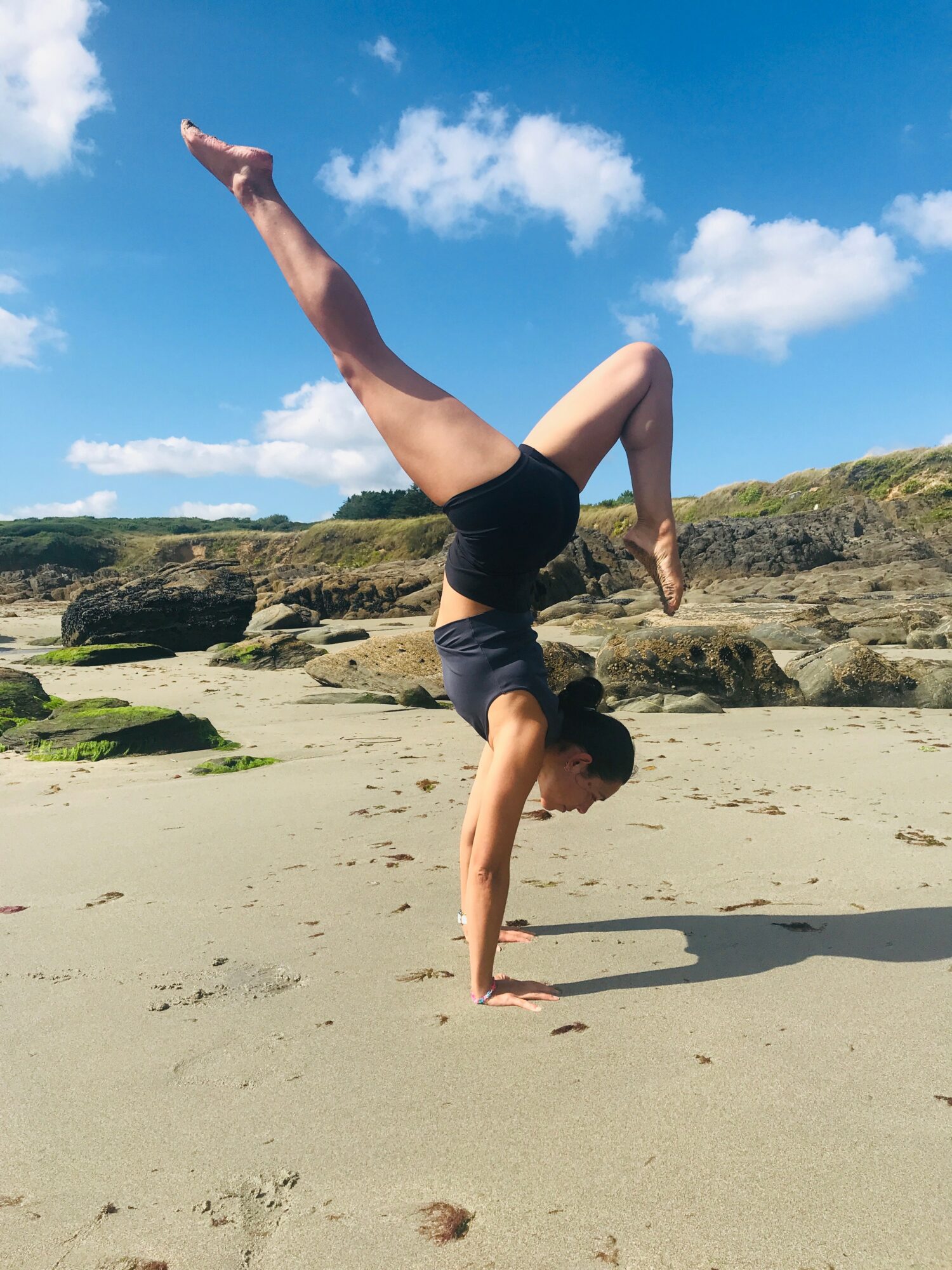 Person performing a handstand on sandy beach with rocks and blue sky in background.