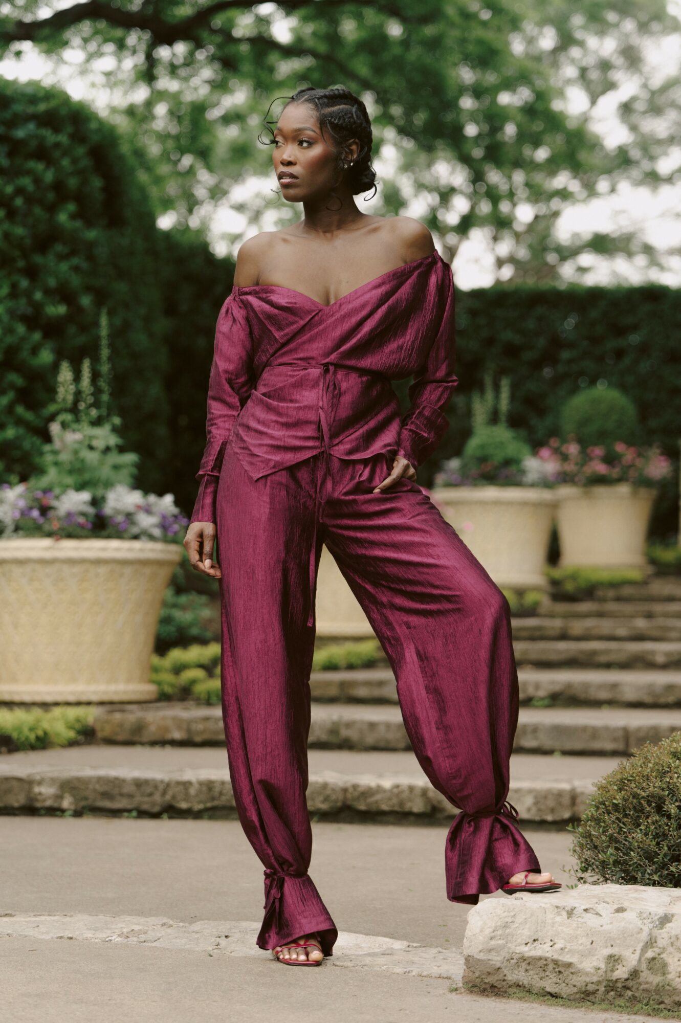 Woman in a maroon off-shoulder outfit standing outdoors with greenery and potted plants in background.