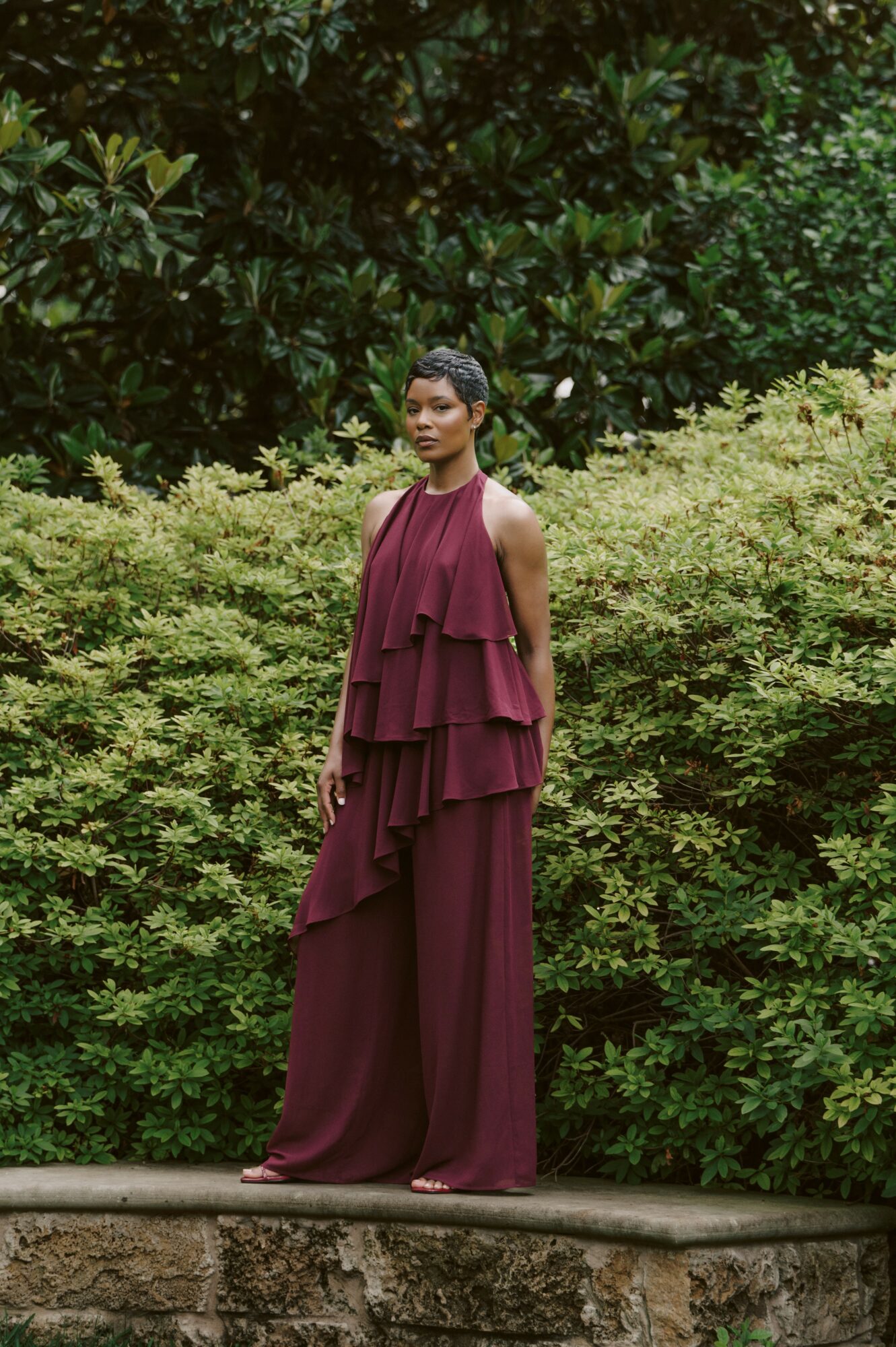 Woman in a long, layered burgundy dress standing on a stone ledge in front of green bushes and trees.