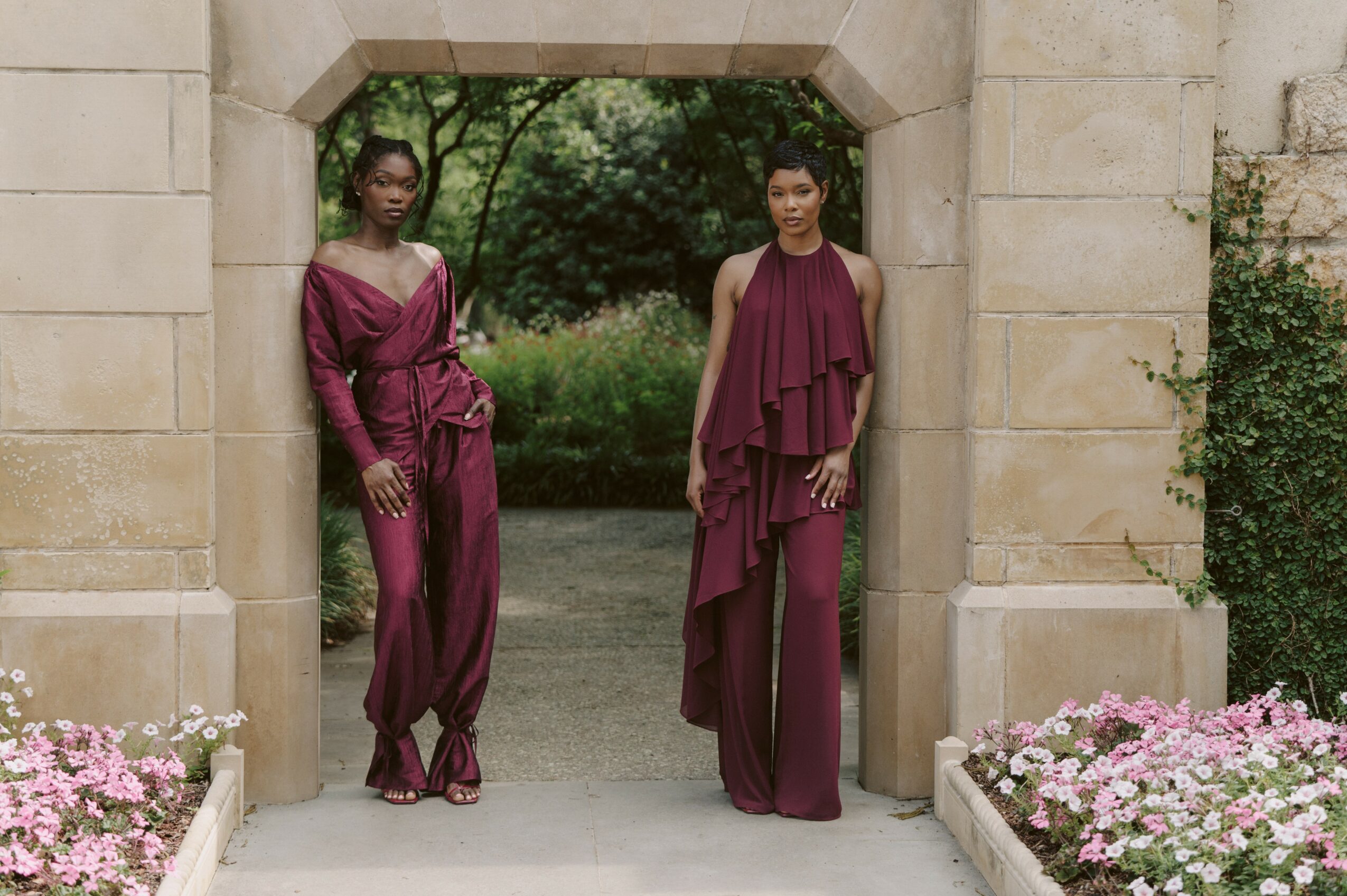 Two women in elegant purple dresses stand in an arched stone doorway with flowers and greenery outside.