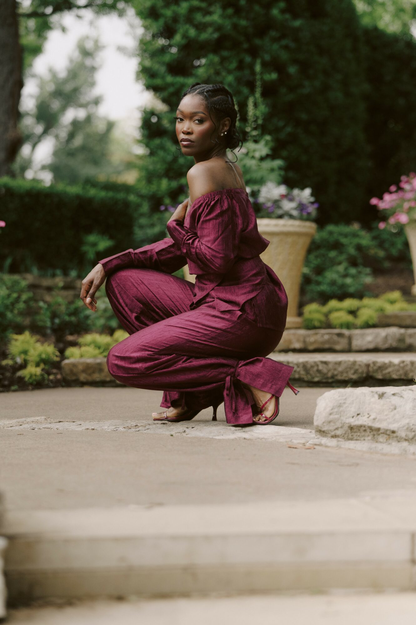 Woman in a maroon off-shoulder jumpsuit crouching outdoors with greenery and flowers in background.