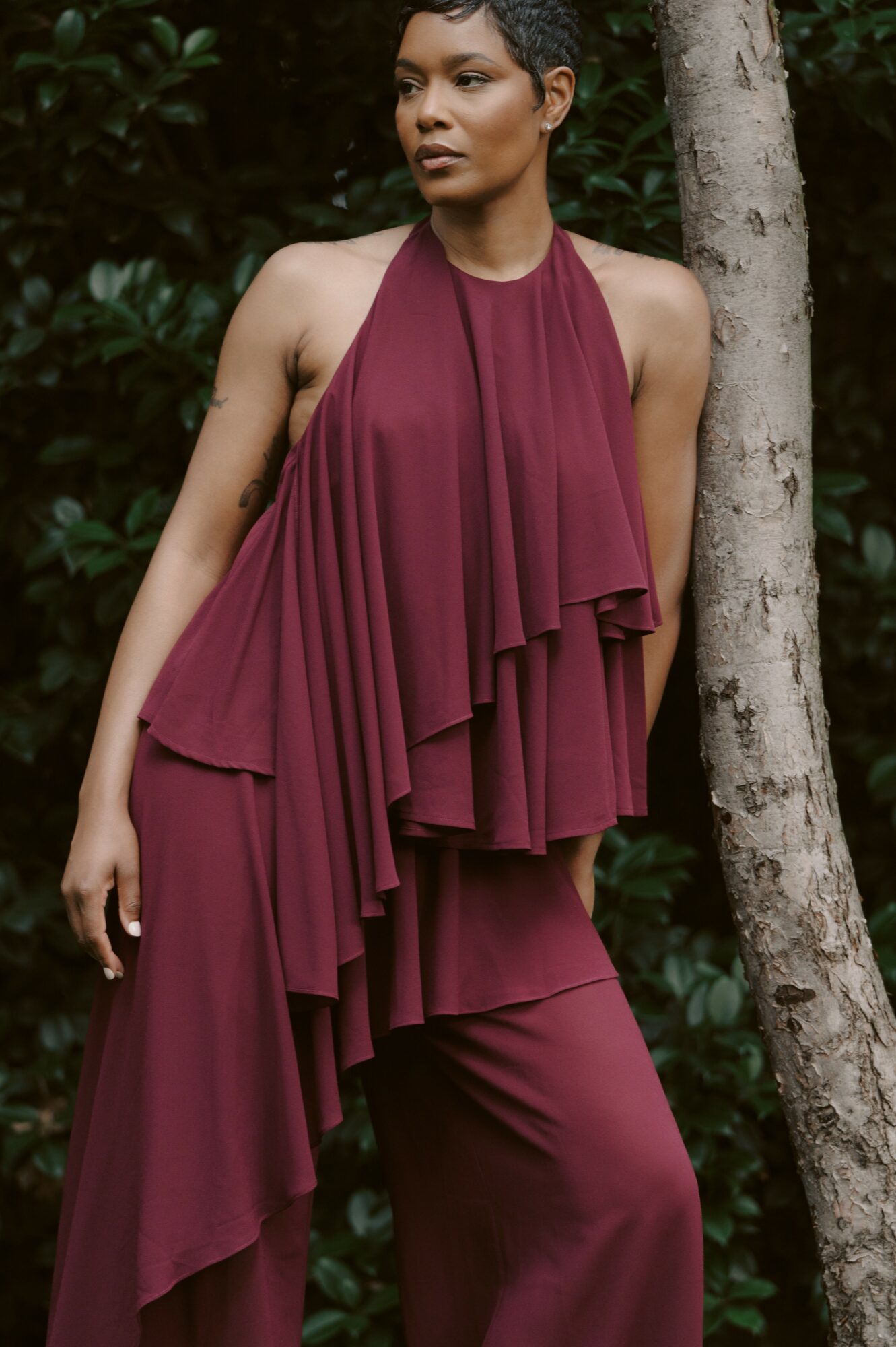 Woman in a sleeveless, burgundy halter dress standing outdoors next to a tree, with greenery in the background.