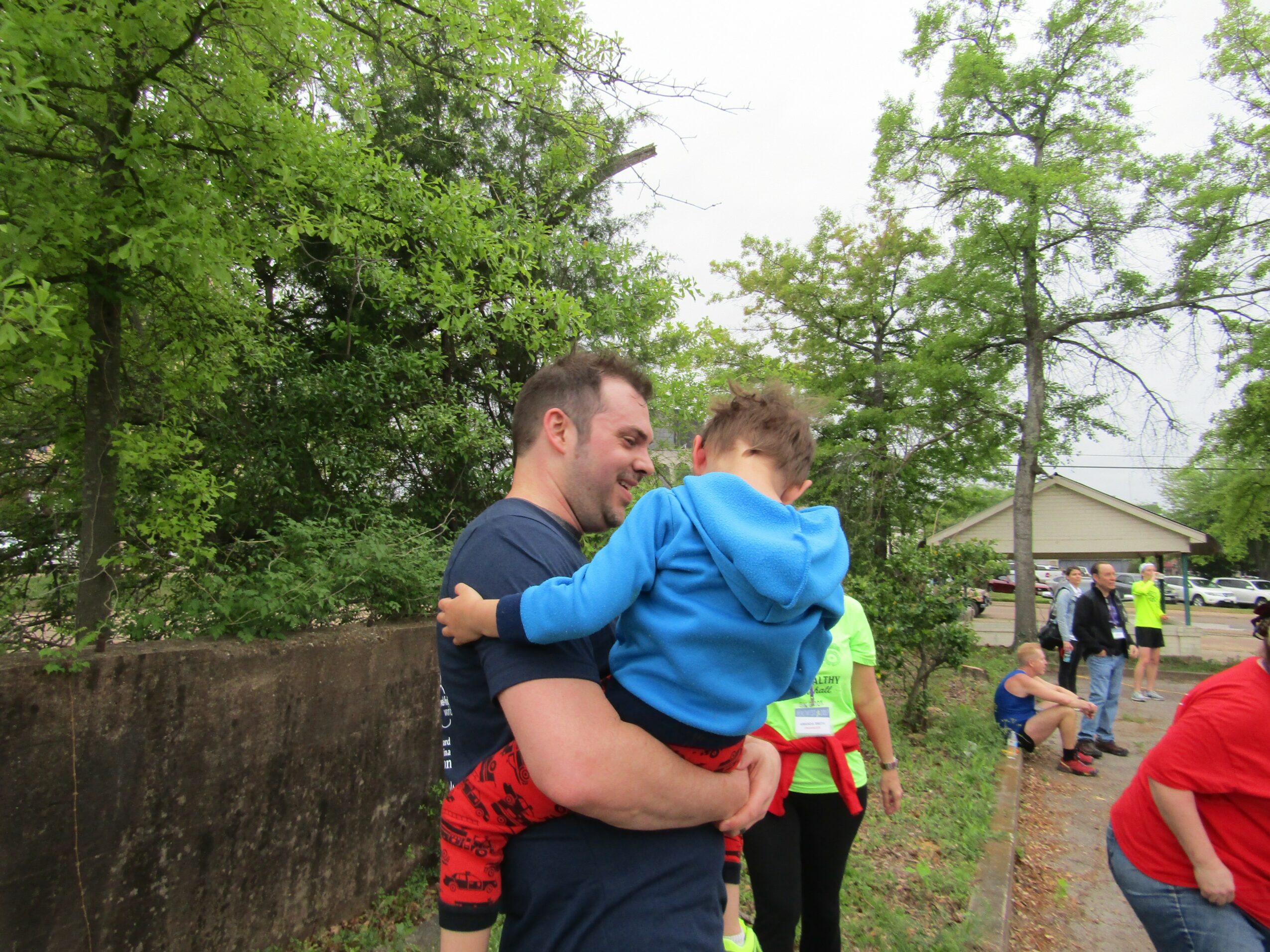 Man holding a child outdoors with trees and people in the background.