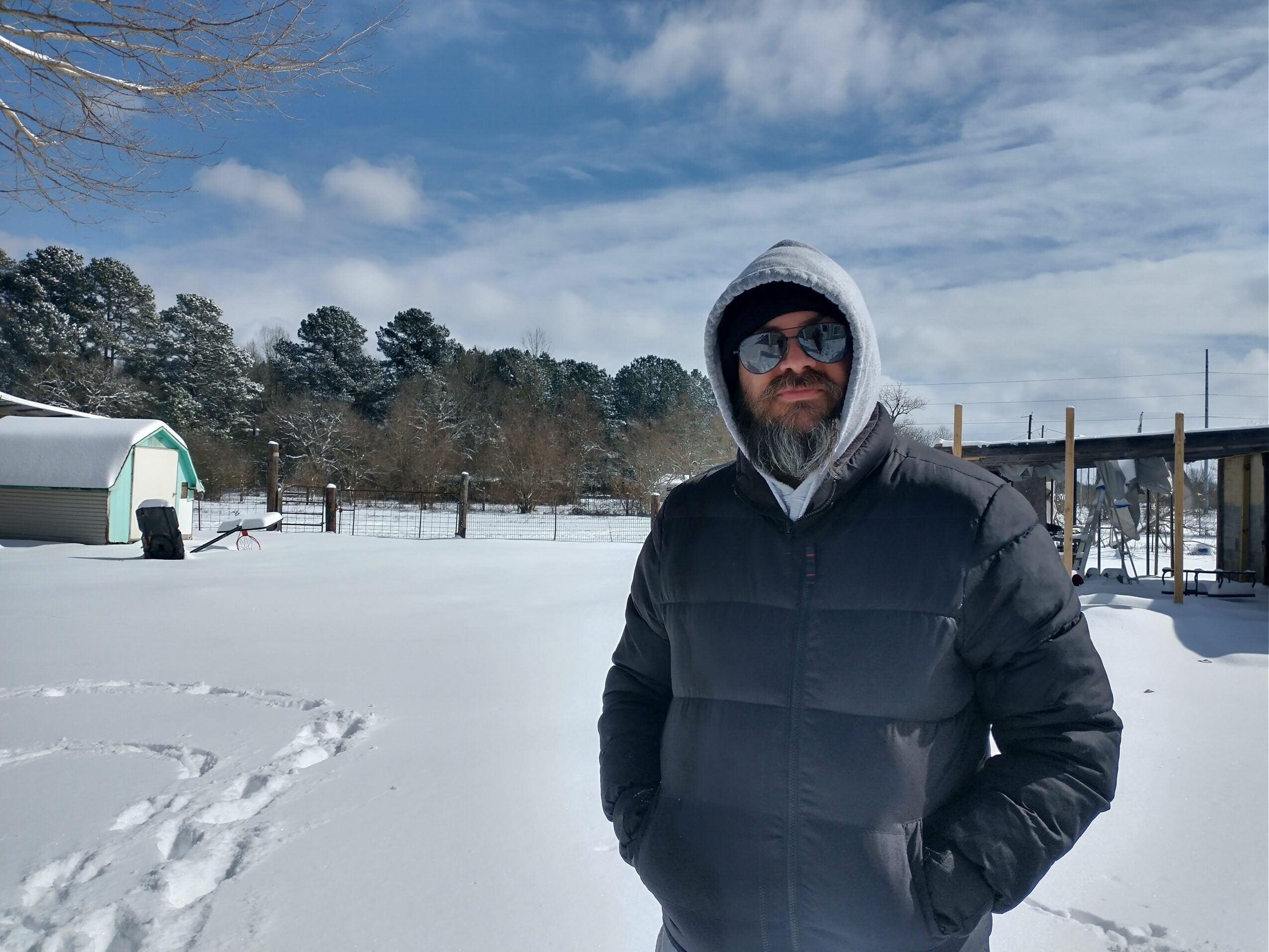 Man in winter coat and sunglasses standing outdoors in snowy landscape with trees and structures in background.