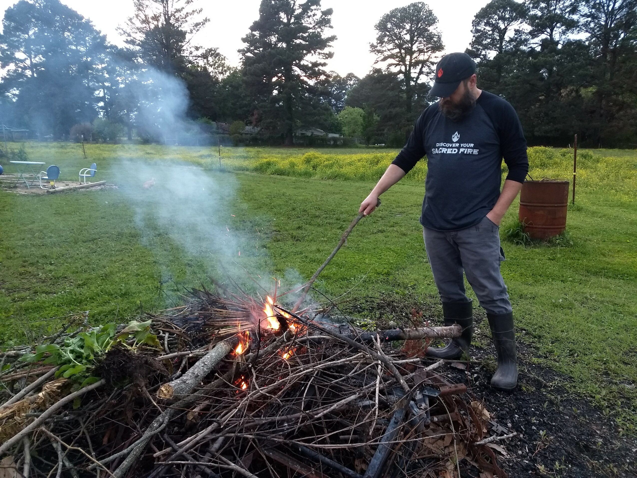 Person tending a small outdoor fire with smoke rising, in a grassy area with trees and a yellow fence in the background.