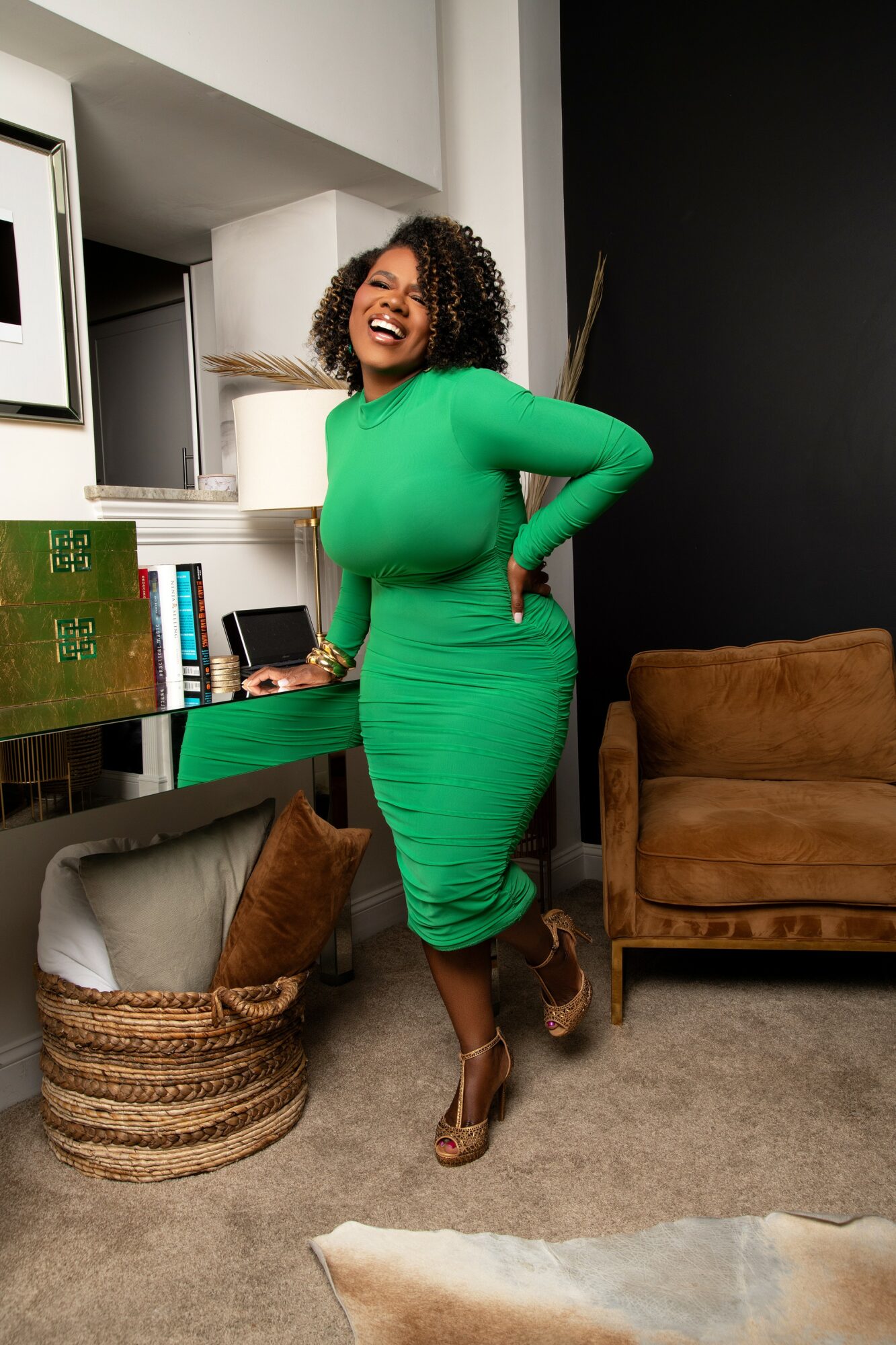 Woman in green dress smiling, standing in a room with a brown chair and a basket, holding a phone, with a black wall behind.