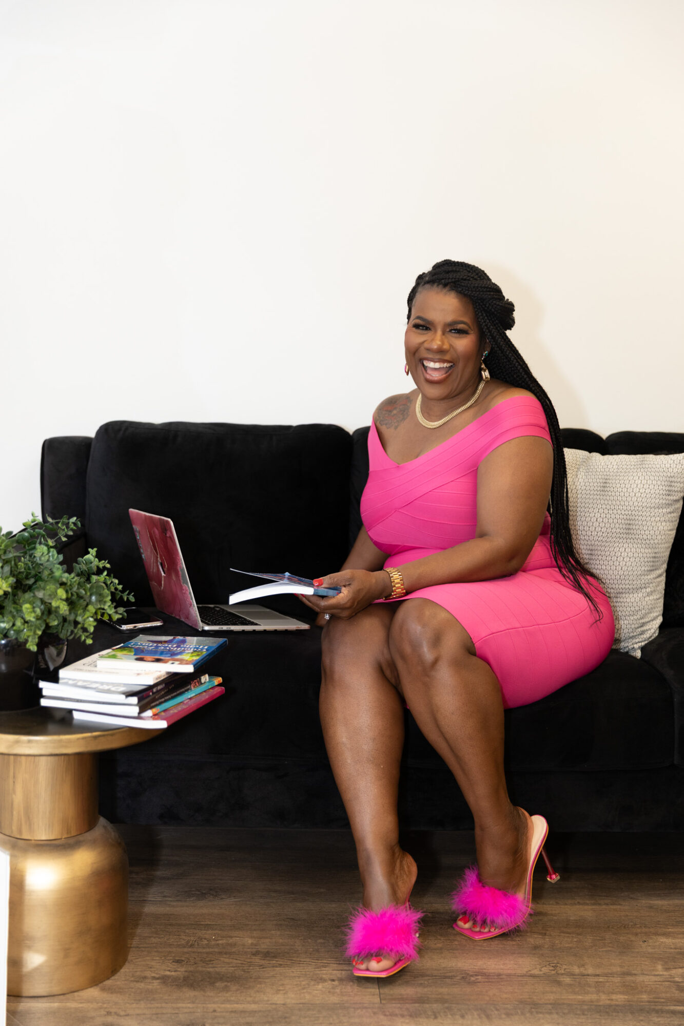 Woman in pink dress sitting on a black sofa, smiling, holding a book, with a laptop nearby, in a room with wooden floor.